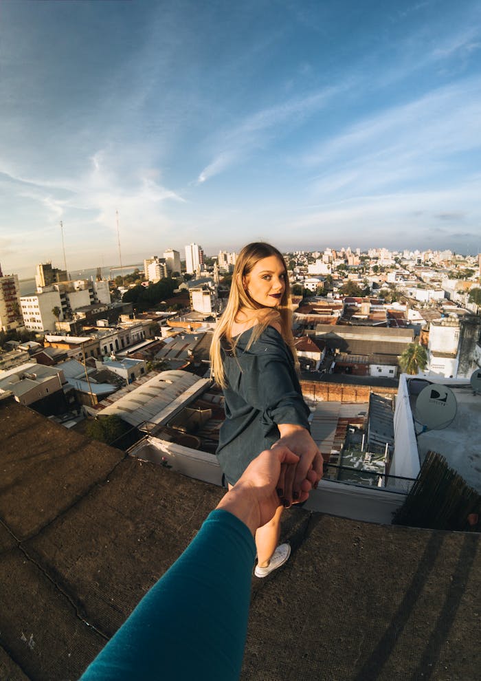 A couple holds hands on a rooftop, overlooking a vibrant cityscape under a blue sky.